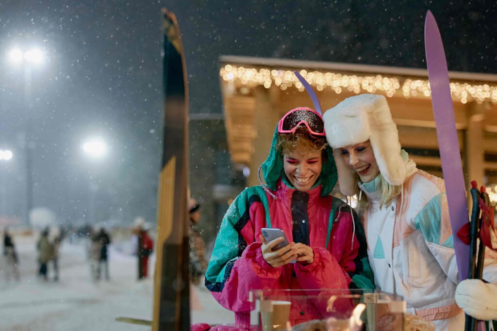 Two female students wearing ski gear laugh at a phone in the Alps